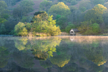 Beautiful Spring colours on trees at Rydal Water in the Lake District, UK. Perfect reflections in misty water with small boathouse. UK landscape photography. - 524676616