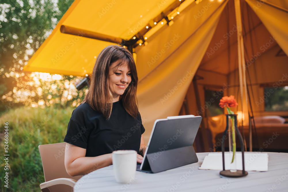 Wall mural smiling woman freelancer using a laptop on a cozy glamping tent in a sunny day. luxury camping tent 