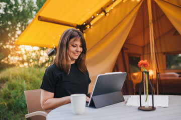 Smiling Woman freelancer using a laptop on a cozy glamping tent in a sunny day. Luxury camping tent...