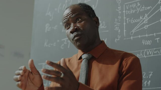 Low angle shot of African American professor standing by chalkboard with math formulas on it and explaining lesson in university