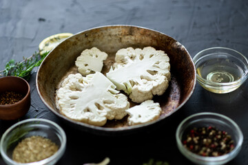 Cauliflower steak cooking. Raw cut cauliflower lie in a frying pan. Olive oil, herbs, various spices nearby. Dark background. Place for text.