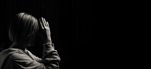 Woman praying with her head bowed with folded palms on a black background. Black and white...