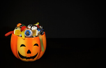 Orange pumpkin jack-o-lantern pot full of assorted candies: traditional Halloween chocolates and jelly worms. Black background with copy space. Trick or treat concept.