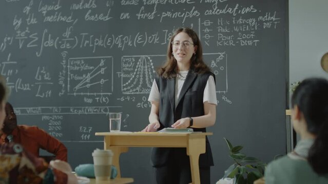 Caucasian female student standing at lectern and talking to group mates during lesson with African American professor