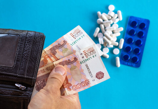 A Russian banknote with a face value of 5000 rubles in the hand against the background of a pills (drugs) on a blue background. Concept of treatment cost, medicine price, health insurance budget