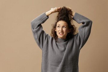 Problematic unruly damaged hair concept. Suffering curly beautiful woman in gray casual sweater with hairbrush comb pulls tangled hair up posing isolated on over beige pastel background. Copy space