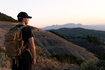 Adolescent boy with a hat and tactical backpack observes the landscape from a mountain at sunset