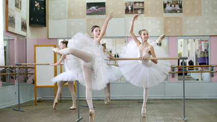in ballet hall, girls in white ballet skirts are engaged at ballet, rehearse attitude, Young ballerinas standing on toes in pointe shoes, raise legs up behind elegantly, at railing in ballet hall