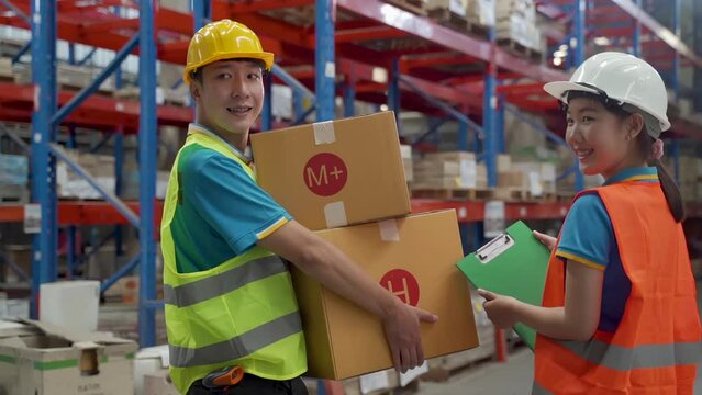 Slow motion, Asian women and men, wearing reflective safety vests and men's safety hats, stand in crates as they prepare to be placed on shelves in a large factory warehouse, smiling happily at work