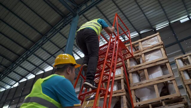 4K, A worker working in a warehouse climbs the red ladder as they go up to check if the goods above are in order. Two men were climbing the stairs and one was keeping an eye on them.