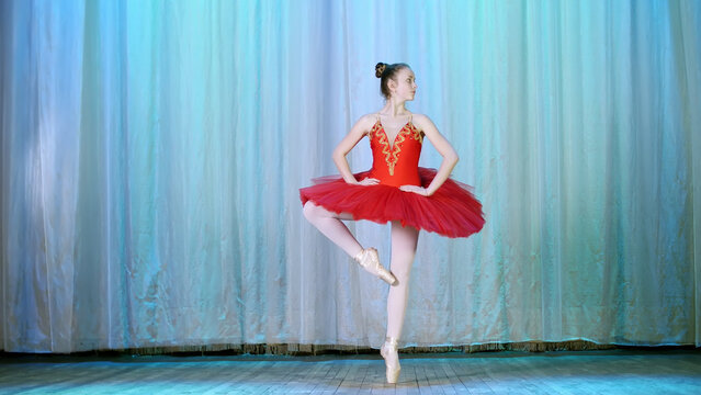 ballet rehearsal, on the stage of the old theater hall. Young ballerina in red ballet tutu and pointe shoes, dances elegantly certain ballet motion, pas courru , tour fouette. High quality photo