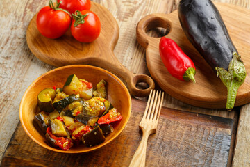 A plate of grilled vegetables next to tomatoes and eggplant on wooden boards next to a wooden fork.