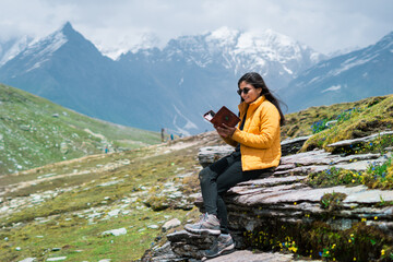 Portrait of Indian teenager girl tourist sitting in front of snowy mountains and reading book. Girl in mountains. Hobby and travel lifestyle concept