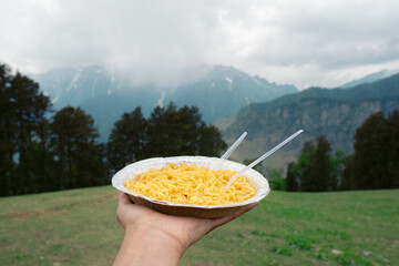 Close up shot of hand holding dish of noodles in background of mountains in Manali, Himachal...