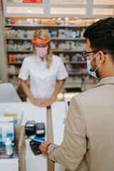 Fototapeta na wymiar Young business man choosing and buying drugs in a drugstore while talking with attractive female pharmacist. She helping him with expert advice. They are wearing protective face masks.