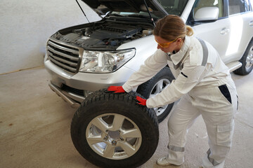 A female car mechanic in a white worker replaces a wheel on a car at a service center. Car components, belts, hoses, labor open hood.