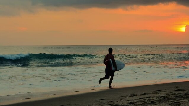 Young male surfer athlete run with surfboard on sandy beach on Bali island sea coast at sunset slow motion video backgroud