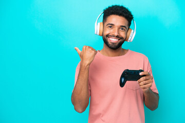 Young brazilian man playing with a video game controller isolated on blue background pointing to the side to present a product - obrazy, fototapety, plakaty