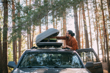 A young man opens the upper tourist trunk of a car in a pine forest on the shore of a lake at...