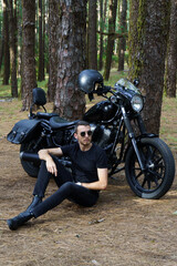 Motero con moto custom descansando junto a su moto en el bosque