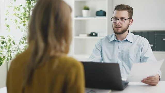 Back view of woman during job interview in bright office. Man asking questions at desk with laptop