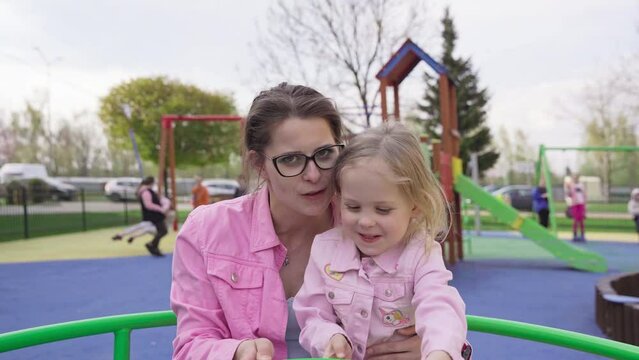A mother and daughter spin on a carousel at a children's playground.