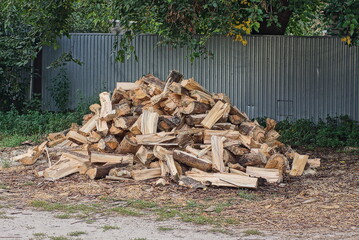 a large pile of chopped brown wooden firewood outdoors in the grass near a gray fence
