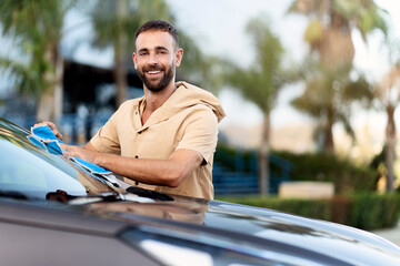 Smiling confident hispanic man cleaning car using micro fiber cloth. Car washing service concept,...