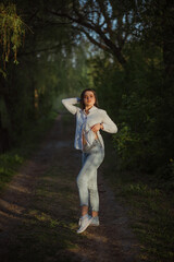 Sexy sensual brunette walks in a summer forest at sunset in a white blouse and jeans