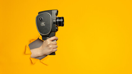 Hand holding small video old retro black camera 8 mm in paper torn hole in yellow background with copy space.
