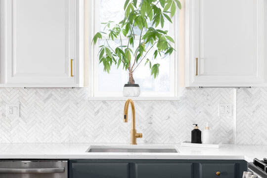 Sink detail shot in a luxury kitchen with herringbone backsplash tiles. white marble countertop, and gold faucet.