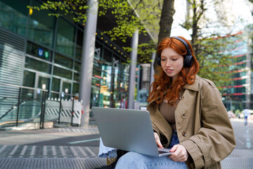 Teen redhead hipster girl student wearing headphones using laptop computer tech device on city street online learning outdoors, elearning outside watching educational webinar sitting in urban park.