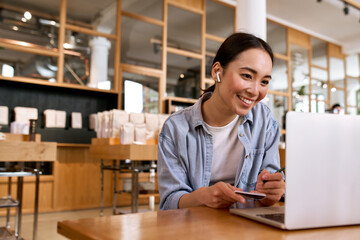 Young smiling Asian woman student using laptop computer wearing earbud, taking notes watching online class elearning webinar training, having hybrid remote video call or virtual work interview.