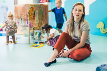 Naklejka premium Young caucasian day-care-center teacher sitting on the floor and looking at the camera while on the background toddlers painting with tempera paints on a cling film wrapped around the wooden shelving