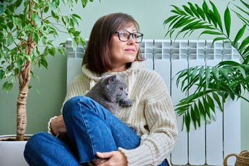 Middle-aged woman in sweater with cat sitting near heating radiator