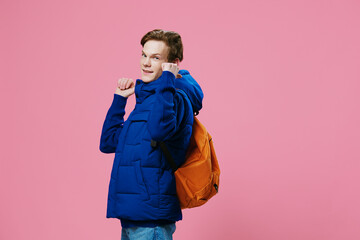 funny funny red-haired guy shows standing sideways to the camera with his finger on a bright backpack on his back while standing in a warm winter jacket on a light blue background