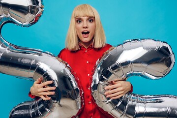 a bright, emotional, funny woman in a red shirt stands on a blue background and holds inflatable balloons in the shape of the number twenty-two in silver color