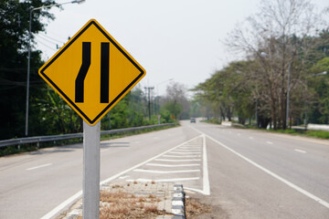 Yellow traffic square shaped left narrow lane sign at rural road Thailand to aware driver know Road narrows on left side ahead. Concept: Warning traffic sign for transportation.           