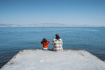 father sitting on a ferry dock with his daughter enjoying the view of beautiful sea with city background