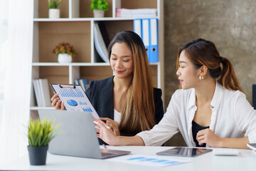 Two professional young Asian business women discuss work, graph, chart using laptop computer for office work.