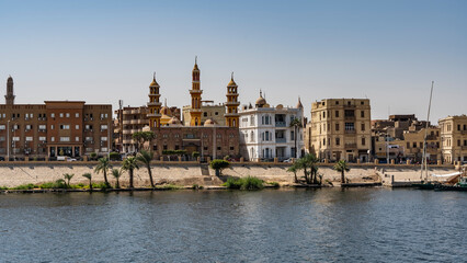 The embankment of the Nile in Egypt. City buildings against a clear blue sky. Palm trees grow at the water's edge. A stone staircase descends to the river