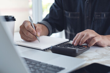 Close up man doing finance at home office, using calculator to calculate expenses on notebook with laptop computer, coffee and mobile phone on office table