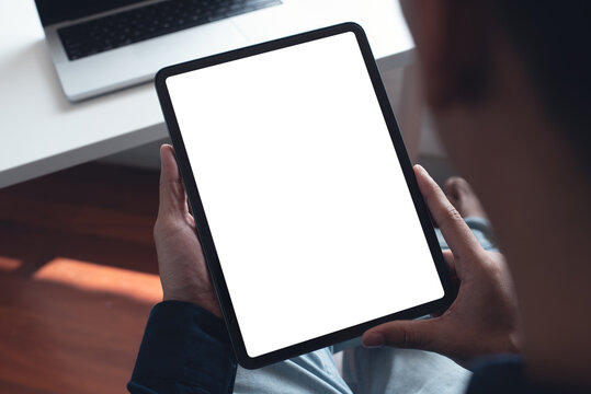 Mockup image of man's hand holding black tablet pc with blank white screen at home office, over shoulder view