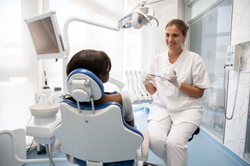 Smiling dentist teaching young female patient to clean teeth with visual aid and toothbrush. African American woman having consultation in dental clinic. Oral hygiene concept