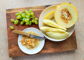 sliced melon with seeds on a white plate, a kitchen knife and green grapes on a wooden board, the concept of fresh fruit and healthy eating