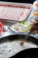 A view hand dipping raw meat slices into the broth bowl, seen with of all the elements of a hot pot restaurant meal.