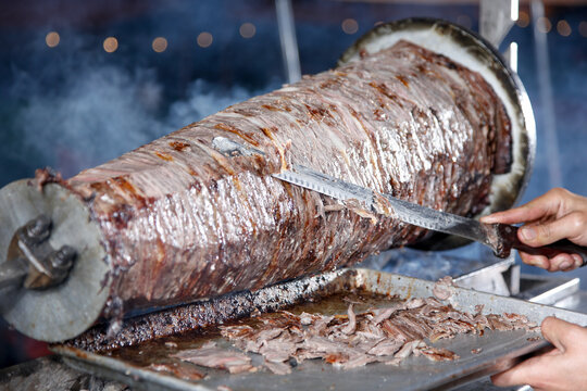 A view of a carving knife cutting trimmings off of beef shawarma cooked on a rotisserie spit.
