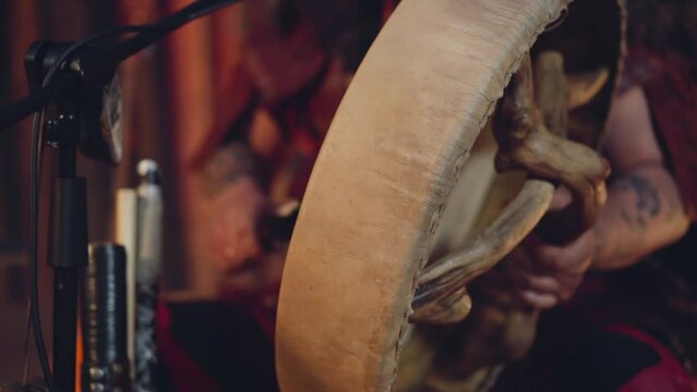 Musician taps shamanic drum with furry stick in ail cabin closeup. Siberian man conducts ancient mystical ritual. Folk music and performance