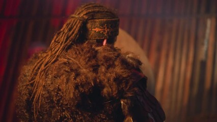 Man with long dreadlocks and fur clothes plays shamanic tambourine at mystical ceremony in ail backside view. Festival of Altai native cultures