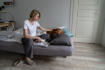 Single middle-aged female looks disappointed at communal bills and strokes favourite dog sitting on bed. Mature blonde woman loses life sense after sudden husband death looking at money debt